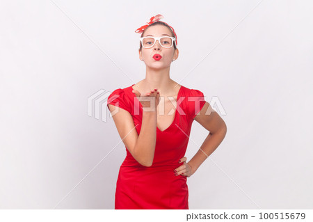 Portrait of pretty lovely cute woman wearing red dress and head band sending air kisses to her boyfriend, keeps han on hip, looking at camera. Indoor studio shot isolated on gray background. 100515679