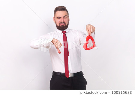 Portrait of unhappy handsome man in white shirt holding little red heart and showing thumb down, dislike gesture, feels disgust of love. Indoor studio shot isolated on gray background. 100515688