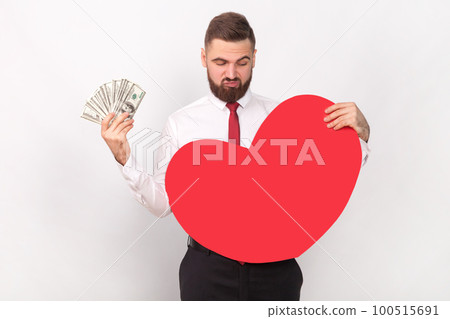 Portrait of young adult bearded man wearing white shirt and tie holding big dollar banknotes, looking at big red heart, choosing money, not love. Indoor shot isolated on gray background. Portrait of young adult bearded man wearing white shirt and tie holding big dollar banknotes, looking at big red heart, choosing money, not love. Indoor shot isolated on gray background. 100515691