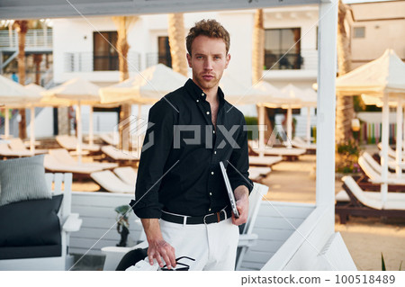 Holds laptop. Young man is outdoors at sunny daytime. Concept of vacation Holds laptop. Young man is outdoors at sunny daytime. Concept of vacation 100518489