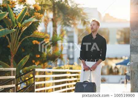 With travel bag. Young man is outdoors at sunny daytime. Concept of vacation 100519900
