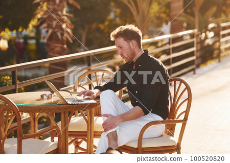 Sits by the table. Young man is outdoors at sunny daytime. Concept of vacation 100520820