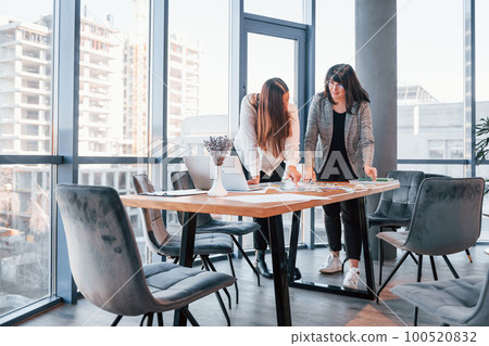 Standing and talking. Two women in formal clothes is indoors in the modern office works together 100520832