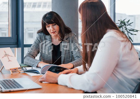 Sitting by the table. Two women in formal clothes is indoors in the modern office works together 100520841