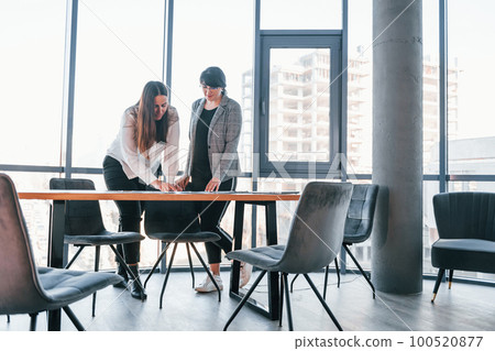 Standing and talking. Two women in formal clothes is indoors in the modern office works together 100520877