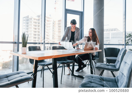 Using laptop. Two women in formal clothes is indoors in the modern office works together 100520880