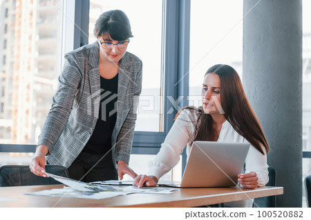 Using laptop. Two women in formal clothes is indoors in the modern office works together 100520882