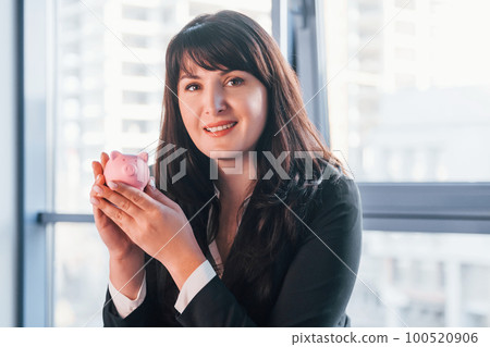 Woman in black formal clothes is indoors in the modern office holds pink piggy bank Woman in black formal clothes is indoors in the modern office holds pink piggy bank 100520906