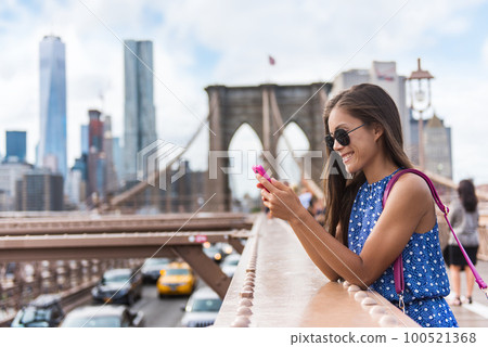 Phone. New York City woman using phone app walking on Brooklyn Bridge by Manhattan city skyline. Young female professional multicultural lady wearing sunglasses, New York City, USA. Phone. New York City woman using phone app walking on Brooklyn Bridge by Manhattan city skyline. Young female professional multicultural lady wearing sunglasses, New York City, USA. 100521368