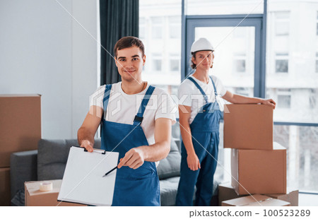 Document to sign. Two young movers in blue uniform working indoors in the room 100523289