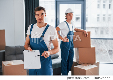 Document to sign. Two young movers in blue uniform working indoors in the room Document to sign. Two young movers in blue uniform working indoors in the room 100523336