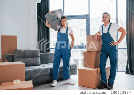 Busy day. Two young movers in blue uniform working indoors in the room 100523554