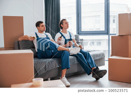 Sitting on the sofa. Tired and taking break. Two young movers in blue uniform working indoors in the room 100523571