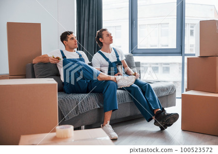 Sitting on the sofa. Tired and taking break. Two young movers in blue uniform working indoors in the room 100523572