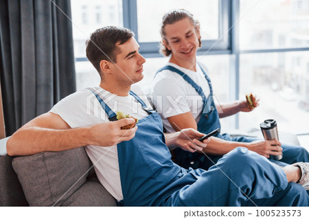Sitting on the sofa. Tired and taking break. Two young movers in blue uniform working indoors in the room 100523573