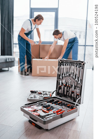 Case with equipment. Two young movers in blue uniform working indoors in the room Case with equipment. Two young movers in blue uniform working indoors in the room 100524198