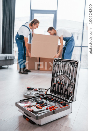 Case with equipment. Two young movers in blue uniform working indoors in the room Case with equipment. Two young movers in blue uniform working indoors in the room 100524200