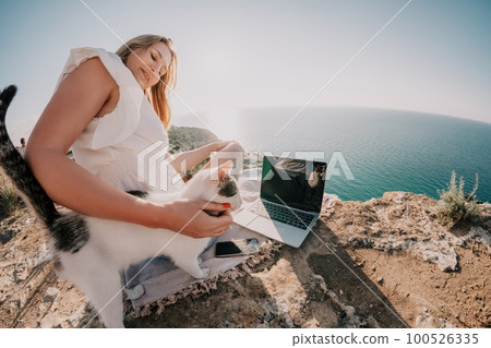 Woman sea laptop. Business woman petting cat and working on laptop by the sea. Close up on hands of pretty lady typing on computer outdoors summer day. Freelance, digital nomad and holidays concept. Woman sea laptop. Business woman petting cat and working on laptop by the sea. Close up on hands of pretty lady typing on computer outdoors summer day. Freelance, digital nomad and holidays concept. 100526335