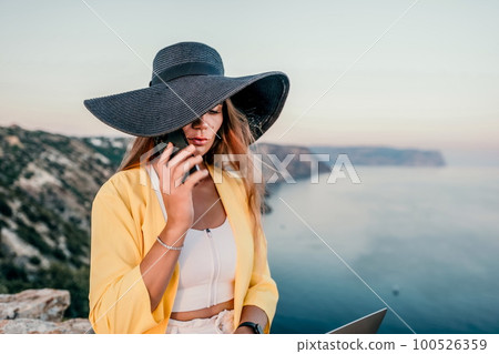 Digital nomad, Business woman working on laptop by the sea. Pretty lady typing on computer by the sea at sunset, makes a business transaction online from a distance. Freelance, remote work on vacation 100526359