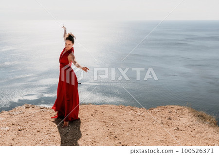 Woman in red dress on sea. Side view a Young beautiful sensual woman in a red long dress posing on a rock high above the sea on sunset. Girl on the nature on blue sky background. Fashion photo. Woman in red dress on sea. Side view a Young beautiful sensual woman in a red long dress posing on a rock high above the sea on sunset. Girl on the nature on blue sky background. Fashion photo. 100526371