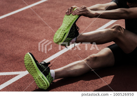 一名男跑步者的下半身在田徑場的跑道上伸展和照顧他的腿 100526950