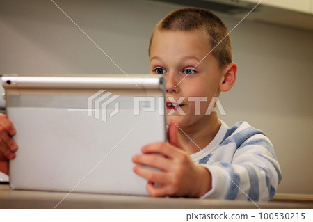Surprised boy 5-10 sitting in the kitchen and looking at the tablet PC. Surprised boy 5-10 sitting in the kitchen and looking at the tablet PC. 100530215
