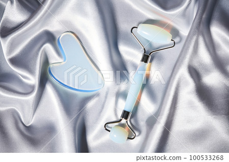 Blue jade face roller and Gua Sha scraping for beauty facial massage therapy on silver textured silk background. Blue jade face roller and Gua Sha scraping for beauty facial massage therapy on silver textured silk background. 100533268