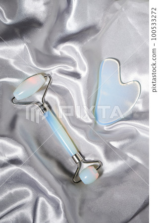 Blue jade face roller and Gua Sha scraping for beauty facial massage therapy on silver textured silk background. Blue jade face roller and Gua Sha scraping for beauty facial massage therapy on silver textured silk background. 100533272