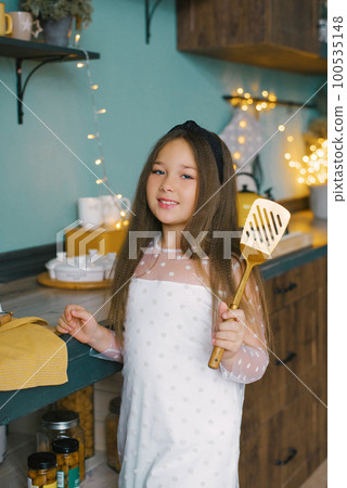 A beautiful girl holds a kitchen spatula in her hands, she wants to cook a festive Christmas dinner against the background of lights in the kitchen A beautiful girl holds a kitchen spatula in her hands, she wants to cook a festive Christmas dinner against the background of lights in the kitchen 100535148