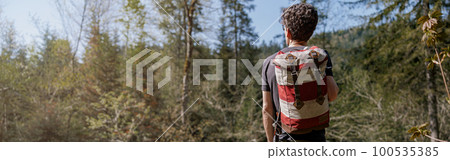 Rear on man with backpack standing on rocks in mountains at wooden bridge over river. 100535385