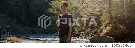 Young male traveler with backpack standing on rocks at river in mountains. Trekking route. Young male traveler with backpack standing on rocks at river in mountains. Trekking route. 100535406