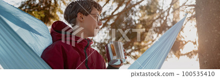 Young woman in glasses sitting in hammock with thermos in forest and enjoying morning. Young woman in glasses sitting in hammock with thermos in forest and enjoying morning. 100535410