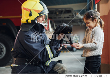 Cute black cat. Happy little girl is with male firefighter in protective uniform Cute black cat. Happy little girl is with male firefighter in protective uniform 100540105