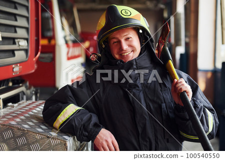 Holds axe in hands. Male firefighter in protective uniform standing near truck 100540550