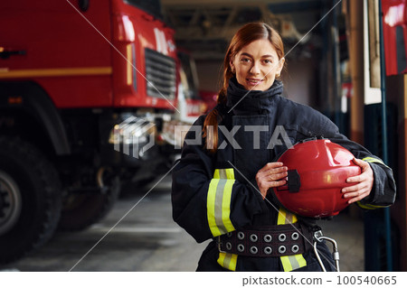 Holds red hat in hands. Female firefighter in protective uniform standing near truck Holds red hat in hands. Female firefighter in protective uniform standing near truck 100540665