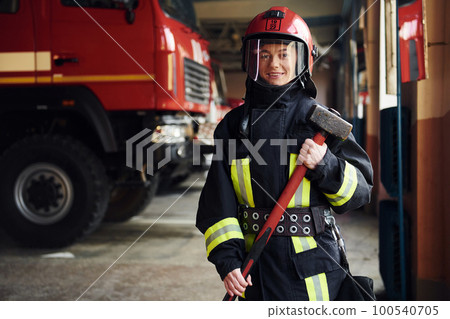 With hammer in hands. Female firefighter in protective uniform standing near truck 100540705