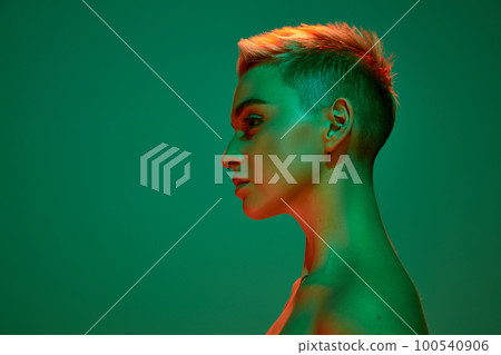 Side view portrait of young blonde girl with short hair and bare shoulders posing against green studio background in orange neon light. Cyberpunk culture Side view portrait of young blonde girl with short hair and bare shoulders posing against green studio background in orange neon light. Cyberpunk culture 100540906