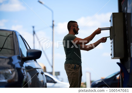 Young man paying for cleaning car on self-service car wash. Silhouette of blue auto and man that going to wash it by active foam. Male person paying for wash service next to car at sunny day. Young man paying for cleaning car on self-service car wash. Silhouette of blue auto and man that going to wash it by active foam. Male person paying for wash service next to car at sunny day. 100547081
