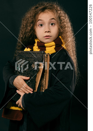 Portrait of a little witch girl with long brown hair dressed in dark coat, holding magic books in her hand, posing on black studio background. Portrait of a little witch girl with long brown hair dressed in dark coat, holding magic books in her hand, posing on black studio background. 100547226