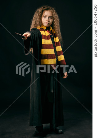 Portrait of a little witch girl with long brown hair dressed in dark coat, holding magic wand in her hand, posing on black studio background. 100547250