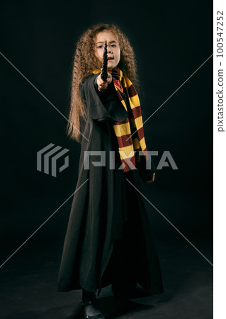 Portrait of a little witch girl with long brown hair dressed in dark coat, holding magic wand in her hand, posing on black studio background. Portrait of a little witch girl with long brown hair dressed in dark coat, holding magic wand in her hand, posing on black studio background. 100547252