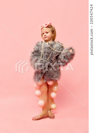 Photo of little stylish blond girl wearing fur coat with sanglasses, touching necklaces, standing and looking away over pink background. Full-length Photo of little stylish blond girl wearing fur coat with sanglasses, touching necklaces, standing and looking away over pink background. Full-length 100547424