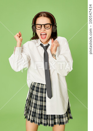 Young, emotional, energetic woman in checkered skirt and white shirt posing, listening to music in headphones against green studio background 100547434