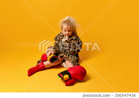 Little angry blond girl wearing boxing gloves and leopard coat sitting and looking away over yellow background Little angry blond girl wearing boxing gloves and leopard coat sitting and looking away over yellow background 100547436