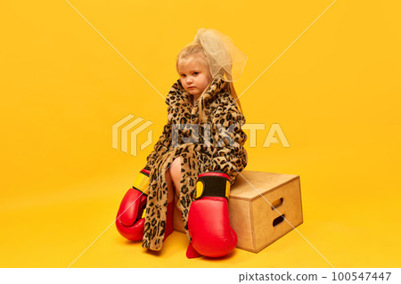 Little upset blond girl wearing boxing gloves and leopard coat sitting on boxes and looking away over yellow background. Little upset blond girl wearing boxing gloves and leopard coat sitting on boxes and looking away over yellow background. 100547447