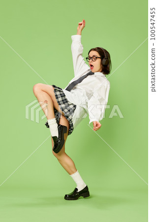 Portrait of young beautiful woman in checkered skirt and white shirt posing, emotionally listening to music in headphones against green studio background Portrait of young beautiful woman in checkered skirt and white shirt posing, emotionally listening to music in headphones against green studio background 100547455