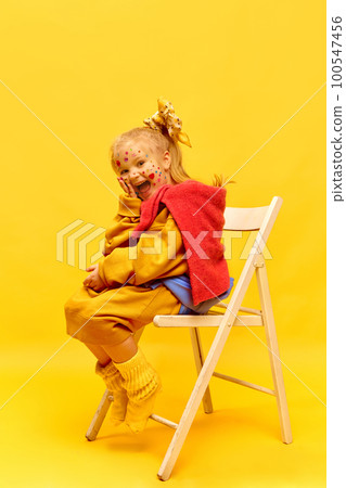 Photo of sweet little girl wearing bow, sitting on chair and laughing at camera over yellow background. Full-length 100547456
