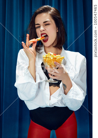 Fast food. Young beautiful brunette woman in white shirt and red tights posing, emotionally eating fries against blue textile background 100547465