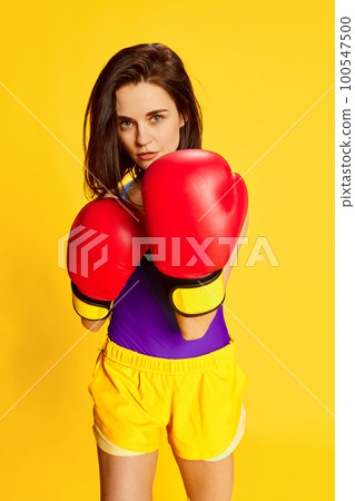 Fitness as hobby. Beautiful young brunette woman in stylish sportswear posing with red boxing gloves against bright yellow studio background. Concept of sport 100547500