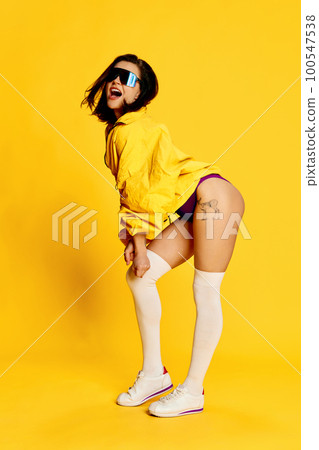 Energy and positivity. Beautiful young brunette woman in stylish sportswear and sunglasses posing against bright yellow studio background. Concept of sport Energy and positivity. Beautiful young brunette woman in stylish sportswear and sunglasses posing against bright yellow studio background. Concept of sport 100547538